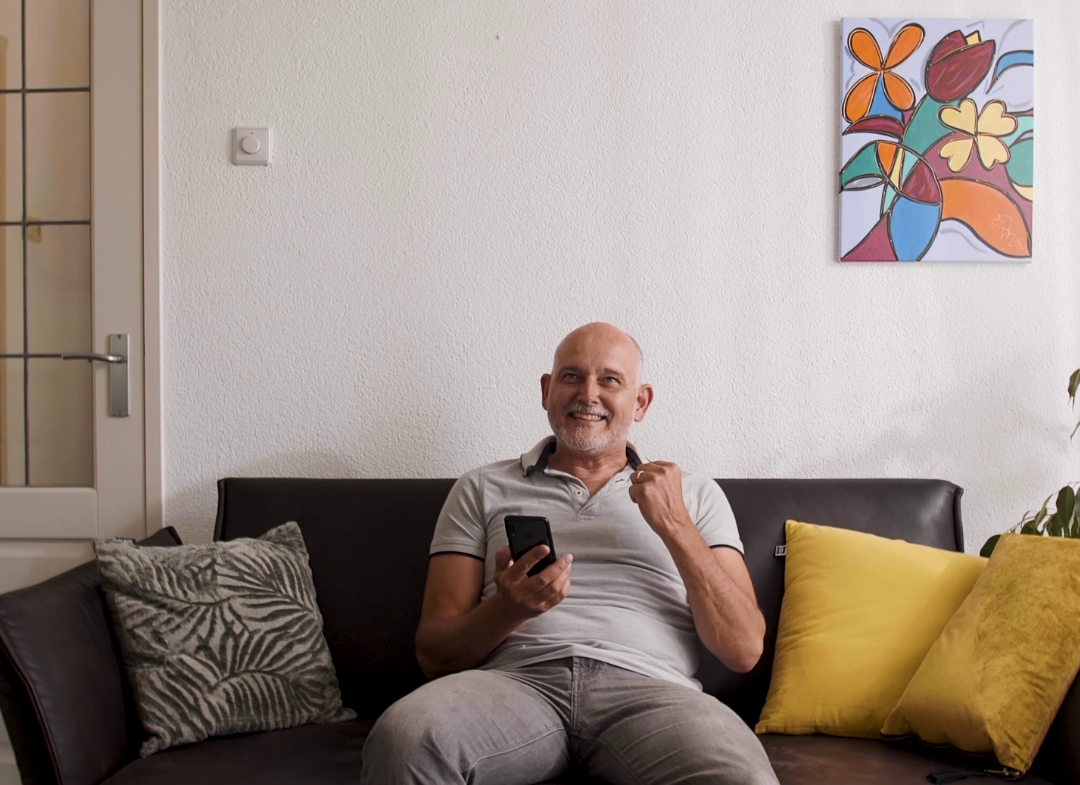 A man in a living room is holding a phone while smiling.