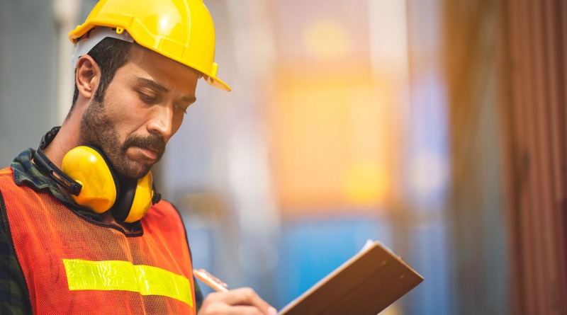 A man wearing a hard hard, and a high visibility jacket writing on the clipboard.
