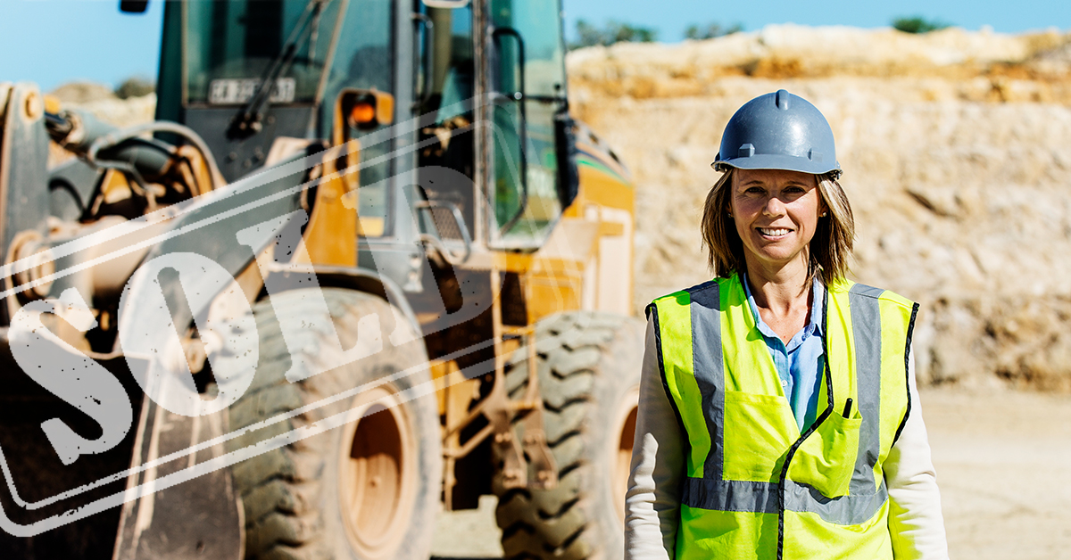 A woman with a hard hat and vest is standing in front of heavy equipment being sold.